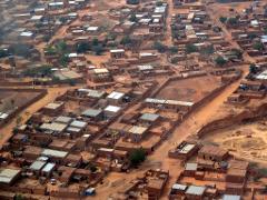 An aerial view captures a dense settlement of mud-brick structures, possibly in an arid region, with simple flat roofs and sandy paths weaving throughout the town.