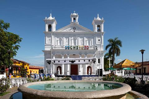 A white church with towers and the word "SUCHITOTO" in colorful letters stands behind a fountain in a town square, with preparations for an event underway.