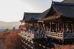 A scenic view of Kiyomizu-dera temple in Kyoto, Japan, showcasing the wooden stage crowded with visitors against the backdrop of colorful autumn foliage and the city skyline.