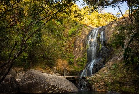 A picturesque waterfall cascades down a rocky cliff, surrounded by lush greenery and a small bridge, creating a serene scene.