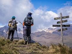 Paramo Trek Day Tour from Medellin Two hikers with backpacks and trekking poles stand on a grassy hill next to a rustic signpost, gazing towards a majestic, snow-capped mountain range under a partly cloudy blue sky.
