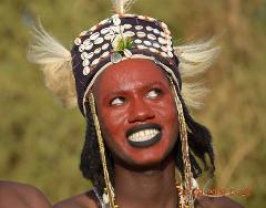 A man with a red face and black lipstick is wearing a headdress decorated with shells and other ornaments and looking upwards with a smile.