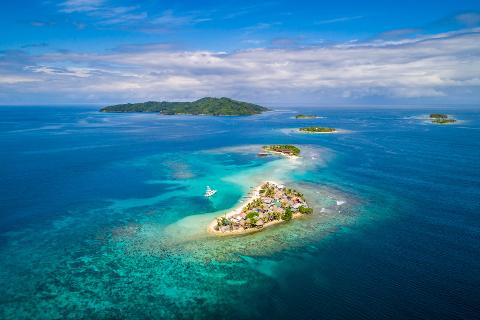 An aerial shot of a small island village surrounded by turquoise waters and other islands in the distance under a partly cloudy sky.