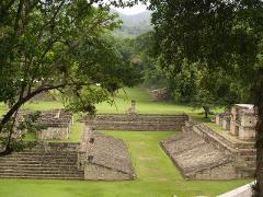 This image shows an aerial view of a Mayan ruin, featuring stone structures, pyramids, and grassy grounds, partly framed by trees.