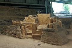 An archeological site reveals ancient mud structures, possibly a temple or residence, with wooden reinforcements, displayed under a modern metal structure.