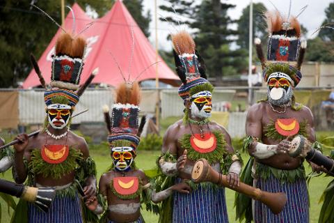 Four colorfully adorned Papua New Guinean men in traditional attire, including face paint, headdresses, and instruments, stand in a row against a backdrop of tents and trees.