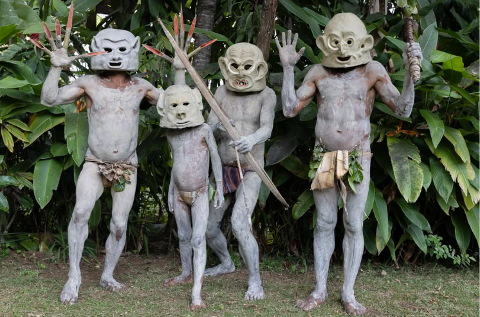 Four men in traditional clay body paint and masks stand with decorated spears and fingers in front of tropical foliage.