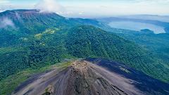 An aerial view of a volcanic landscape with a cone-shaped volcano in the foreground, lush green mountains, and a lake in the distance.