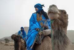 A man in blue robes and turban rides a camel in a desert landscape, with another man and camel in the background.