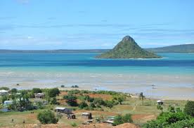 The image showcases a scenic coastal landscape with a verdant, cone-shaped island in the turquoise water, a sandy shore, and a village with scattered buildings in the foreground.