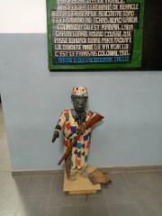This image shows a statue of an African man in a colorful coat holding a rifle, standing beneath a sign with French text about colonial history.