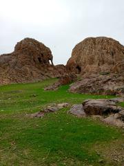A landscape with two large brown rock formations flanking a green grassy area under a cloudy sky. The left rock has a distinct hole resembling an elephant's trunk.