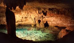 A cavern with stalactites and stalagmites over clear water with rocks visible on the bottom.