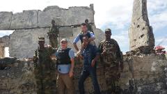 A group of seven people, including four armed soldiers in camouflage, pose in front of a ruined building with a partly cloudy sky in the background.