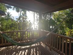 A green and blue hammock hangs on a wooden porch overlooking lush greenery with the sun shining through the trees.