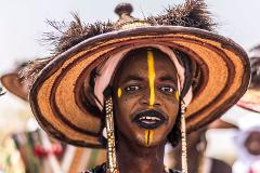 A man in a wide-brimmed hat adorned with feathers and traditional yellow face paint smiles at the camera.