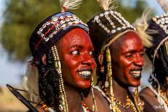 Two men with red painted skin are dressed in traditional headdresses and colorful jewelry, smiling with black painted lips in an outdoor setting.