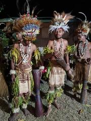 Three men in traditional Papua New Guinean attire, including headdresses, shell necklaces, and woven skirts with foliage, stand with drums or percussion instruments.