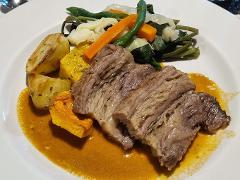 A plate of braised meat in sauce is presented alongside roasted potatoes, squash, and a medley of steamed vegetables including green beans, carrots, and cabbage.