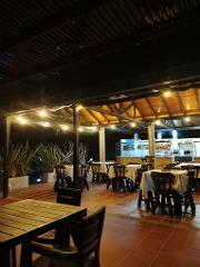 The image shows an outdoor dining area at night, illuminated by string lights, with tables and chairs on a tiled floor, and a serving area in the background.