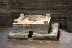 An ancient, weathered, stone-like structure with a central depression stands in an earth-toned chamber with layered soil walls.