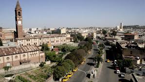 Here's a description of the image:A high-angle shot of Asmara, Eritrea, shows a city with Italian colonial architecture, including a prominent clock tower and a church, set against a clear sky.