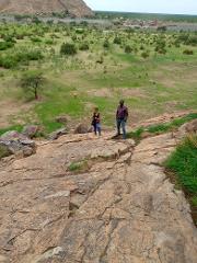 A man and a woman stand on a rocky outcrop overlooking a grassy plain dotted with trees and a distant hill under a cloudy sky.