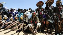 A group of men in traditional attire, some kneeling and others standing, are participating in a cultural festival. They wear hats and face paint.