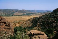 A vast, arid landscape unfolds under a clear sky, with rocky hills in the foreground giving way to a flat plain and distant cliffs.