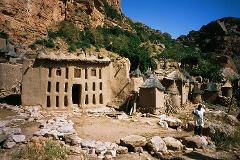 A village of earthen buildings with thatched roofs nestles against a rocky cliff, with a man standing in the foreground.