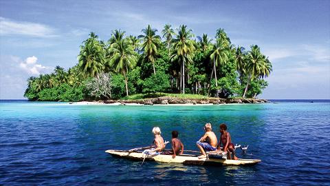 A group of children are in a small boat in the clear blue waters in front of a tropical island covered with lush green trees.