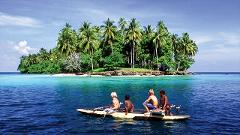 A group of children are in a small boat in the clear blue waters in front of a tropical island covered with lush green trees.