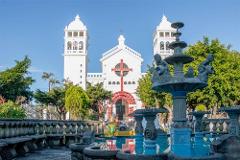 A white church with two towers is seen behind a fountain and ornate benches in a sunny, outdoor setting.