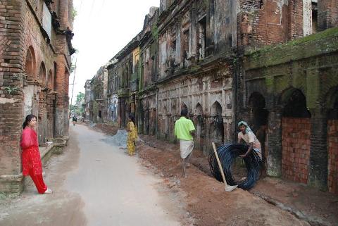 3-Day Dhaka Heritage & Culture Tour — Old Palaces, Lost Cities & Zamindari Mansions A street lined with ancient, weathered buildings shows people working on repairs and walking, with a woman in a red outfit leaning against a brick wall.