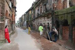 3-Day Dhaka Heritage & Culture Tour — Old Palaces, Lost Cities & Zamindari Mansions A street lined with ancient, weathered buildings shows people working on repairs and walking, with a woman in a red outfit leaning against a brick wall.