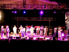 A music band performs on a stage with "FESTIVAL SUR LE NIGER" sign under the stage lights, viewed from a crowd.