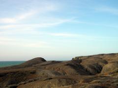 A rugged, arid landscape of brown hills and sparse vegetation meets the sea under a pale blue sky.