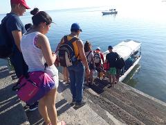 A group of people are boarding a white boat on the water from a concrete pier with steps, some carrying bags and water bottles.