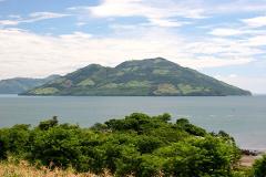 A scenic view of a lush, green island rising from the tranquil waters, under a partly cloudy blue sky, captured from a vantage point amidst vibrant greenery.