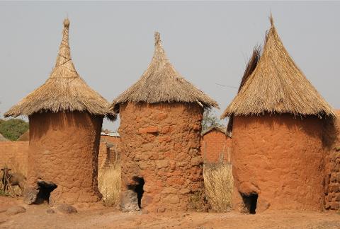 Three traditional mud-brick granaries with thatched conical roofs stand in a dusty landscape under a hazy sky.