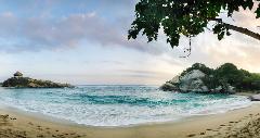 Parque Nacional Natural Tayrona Tour de 2 días desde Cartagena A scenic beach panorama shows turquoise waves meeting a sandy shore, with rocky outcrops on each side, one topped with a small hut, under a partially cloudy sky and framed by overhanging green leaves.