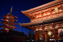 A nighttime view of the Senso-ji Temple in Tokyo, featuring the five-storied pagoda and the Kaminarimon gate illuminated in red and white against a deep blue sky.