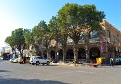 A row of cars parks along a tree-lined street in front of a historic building with arched facades, with people walking nearby under a clear blue sky.