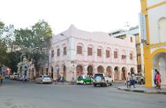 A bustling street scene in a town with a pink building named "Kiosque Hayat" featuring arched windows and columns, next to a yellow and white building marked "LABO AFFI."