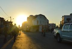 A street scene at sunset, with silhouettes of people walking, buildings lining the road, and a pickup truck on the right.