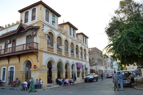 A street scene in a historical town with European-style architecture, people sitting and standing around, and vehicles parked along the street.