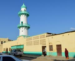 A white and green minaret rises above a yellow building, with two women standing outside the door.