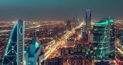 A nighttime view of the illuminated Riyadh skyline, featuring prominent skyscrapers and brightly lit streets.