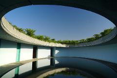 A circular courtyard with a reflecting pool beneath an open concrete roof lined with greenery under a clear blue sky.