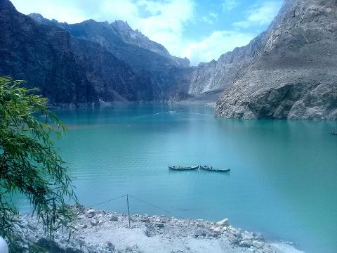 A scenic view of a turquoise lake surrounded by towering mountains, with two boats gently floating on the water, framed by lush greenery in the foreground, all under a cloudy blue sky.